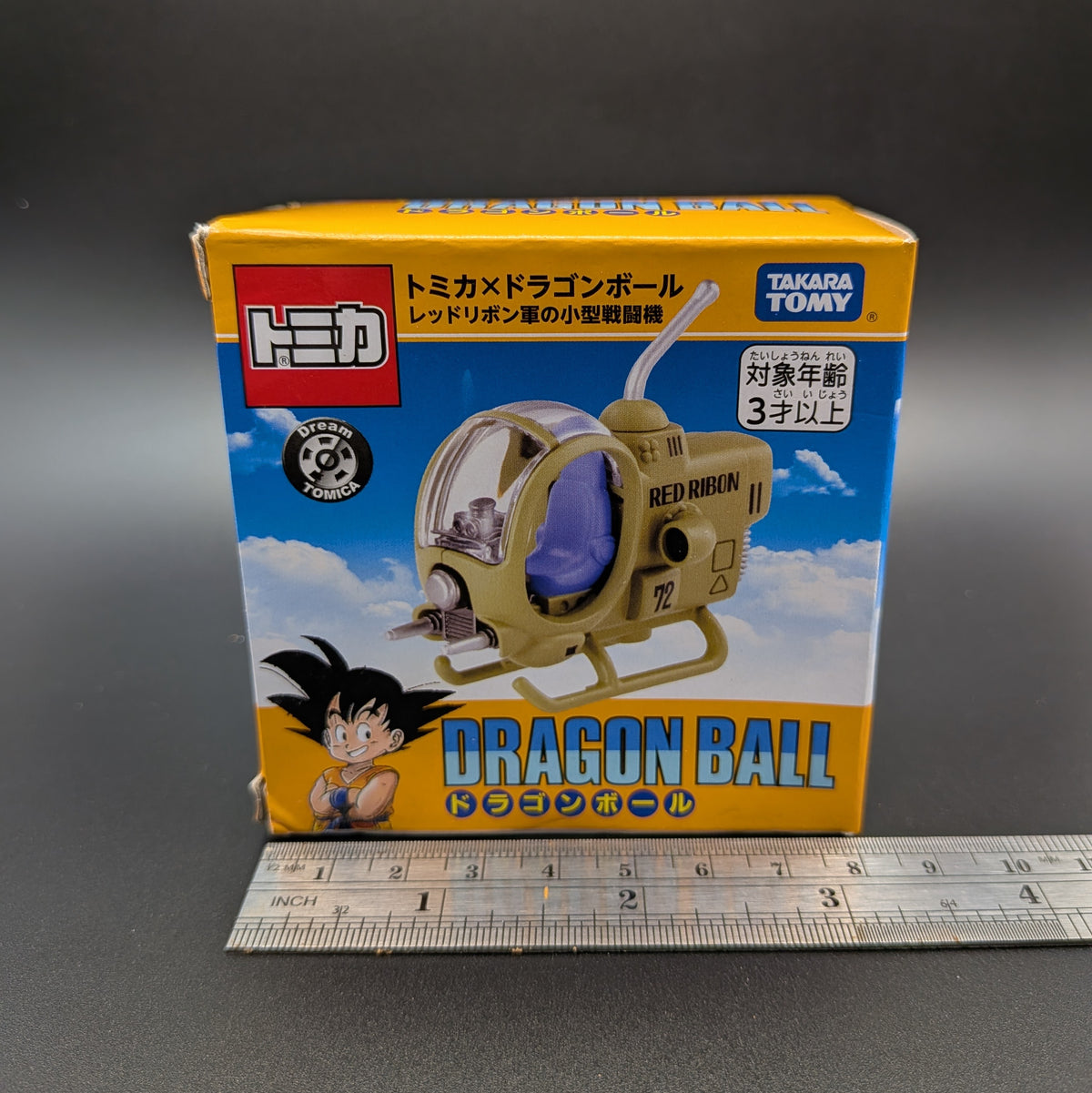 Tomy Tomica Dragon Ball Red Ribbon Helicopter toy packaging box shown upright on a dark gray surface, with a metal ruler placed in front for scale. The box features Dragon Ball and Goku artwork and the Takara Tomy and Tomica branding.