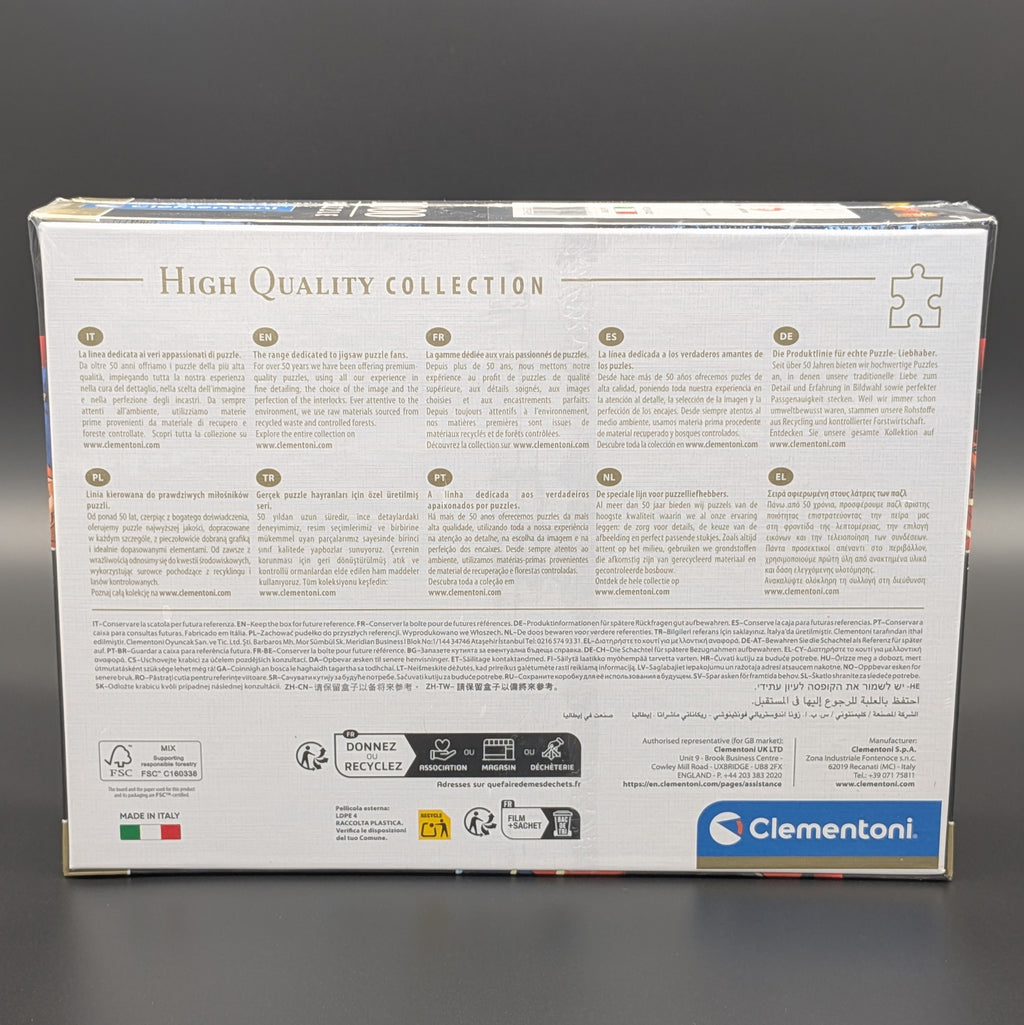The back of a Clementoni jigsaw puzzle box, showing product information, recycling icons, and a blue Clementoni logo on a white background, photographed against a dark gray backdrop.