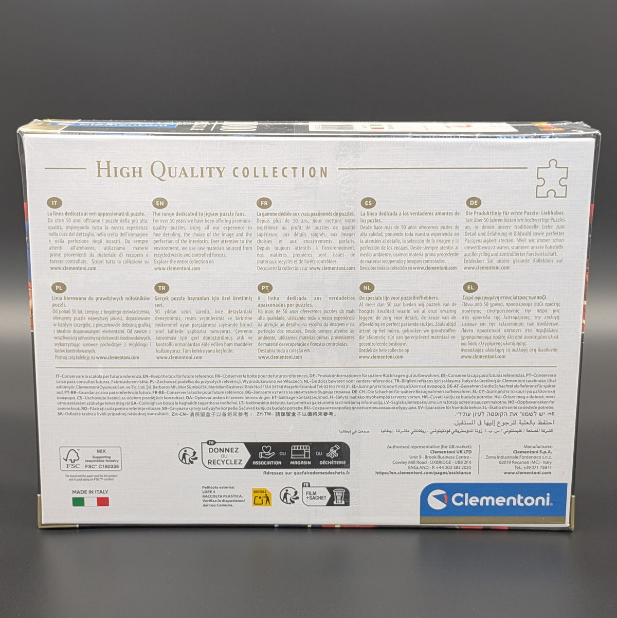 The back of a Clementoni jigsaw puzzle box, showing product information, recycling icons, and a blue Clementoni logo on a white background, photographed against a dark gray backdrop.
