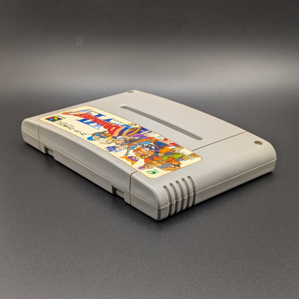 Dragon Quest 6 video game cartridge for Nintendo's Super Famicom (SFC) Entertainment System, shown laying on its back with the label facing upwards. Showing the left hand side and top edge. It sits on a dark grey background.
