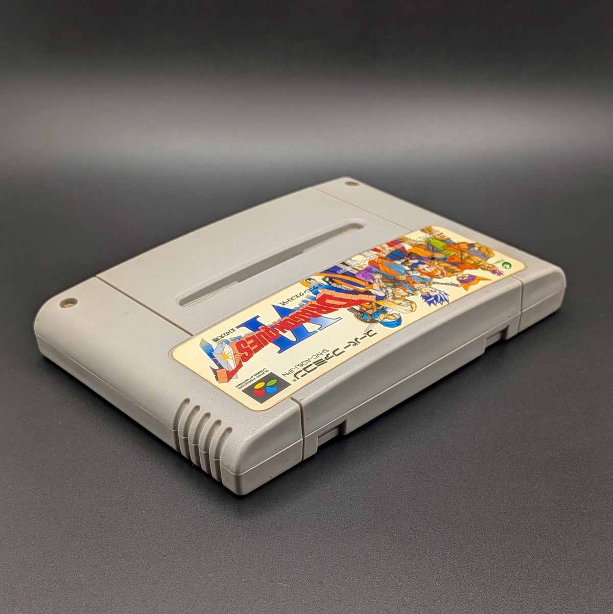 Dragon Quest 6 video game cartridge for Nintendo's Super Famicom (SFC) Entertainment System, shown laying on its back with the label facing upwards. Showing the right hand side and top edge. It sits on a dark grey background.