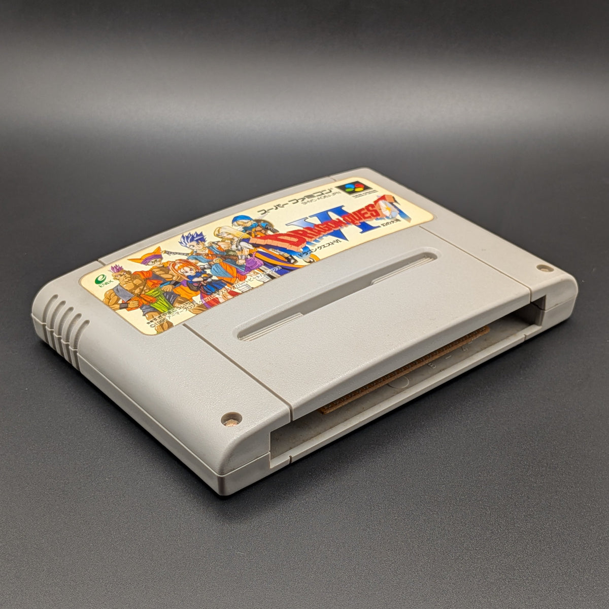 Dragon Quest 6 video game cartridge for Nintendo's Super Famicom (SFC) Entertainment System, shown laying on its back with the label facing upwards. Showing the left hand side and bottom slot. It sits on a dark grey background.