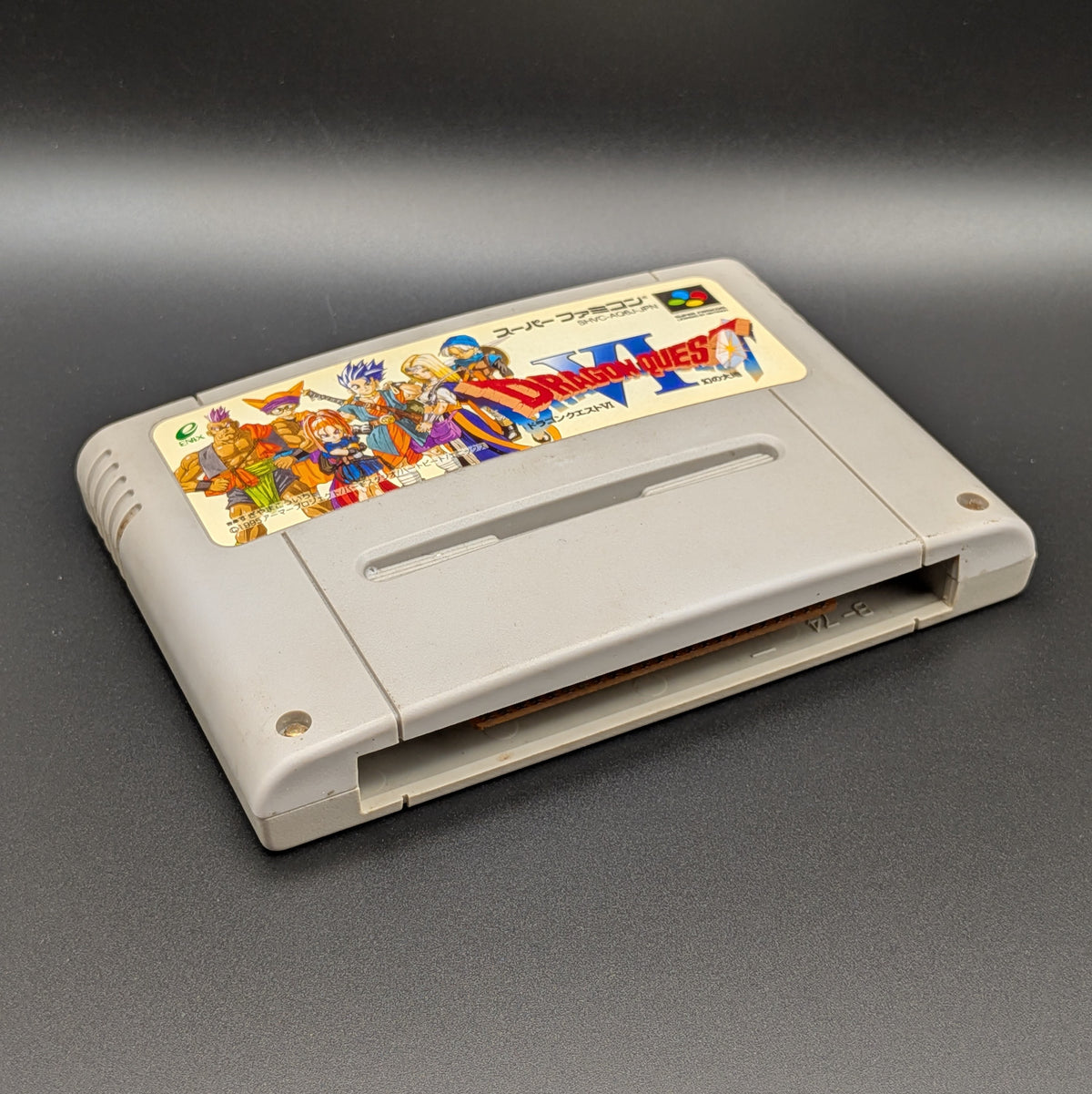 Dragon Quest VI video game cartridge for Nintendo's Super Famicom (SFC) Entertainment System, shown laying on its back with the label facing upwards. Showing the left hand side and bottom slot. It sits on a dark grey background