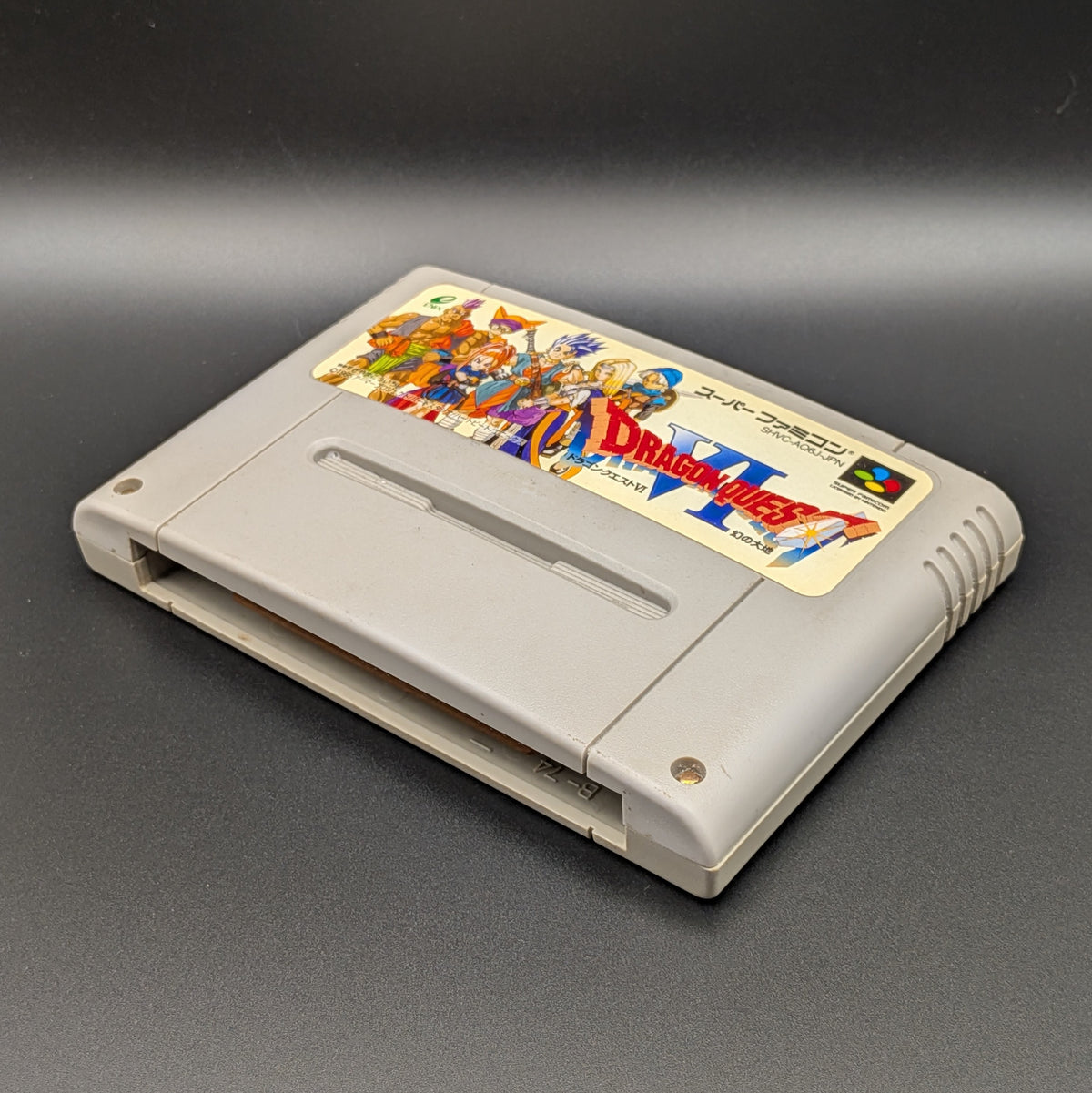 Dragon Quest VI video game cartridge for Nintendo's Super Famicom (SFC) Entertainment System, shown laying on its back with the label facing upwards. Showing the right hand side and bottom slot. It sits on a dark grey background