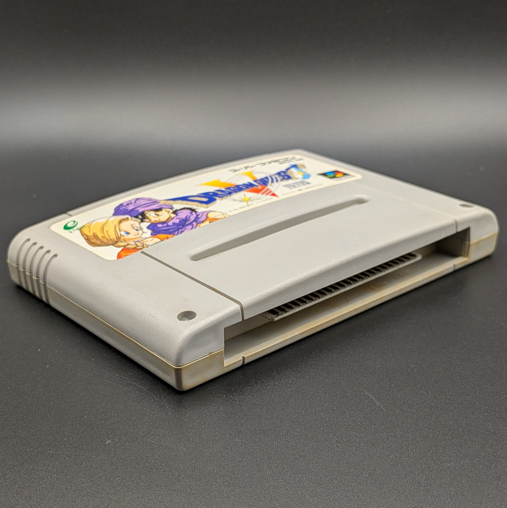Dragon Quest V video game cartridge for Nintendo's Super Famicom (SFC) Entertainment System, shown laying on its back with the label facing upwards. Showing the left hand side and bottom slot. It sits on a dark grey background.