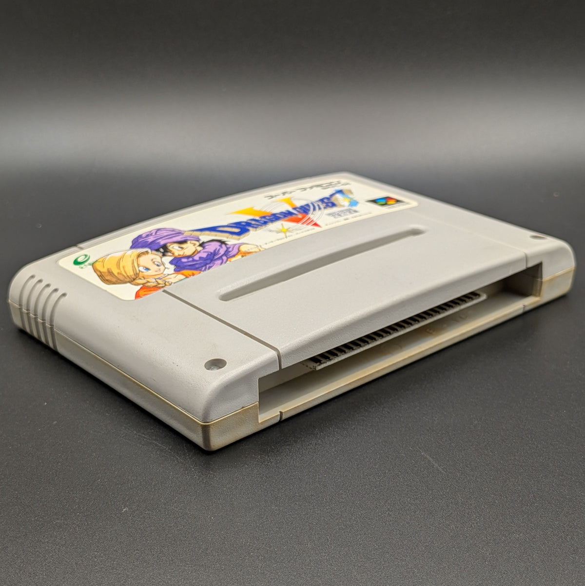 Dragon Quest V video game cartridge for Nintendo's Super Famicom (SFC) Entertainment System, shown laying on its back with the label facing upwards. Showing the left hand side and bottom slot. It sits on a dark grey background.