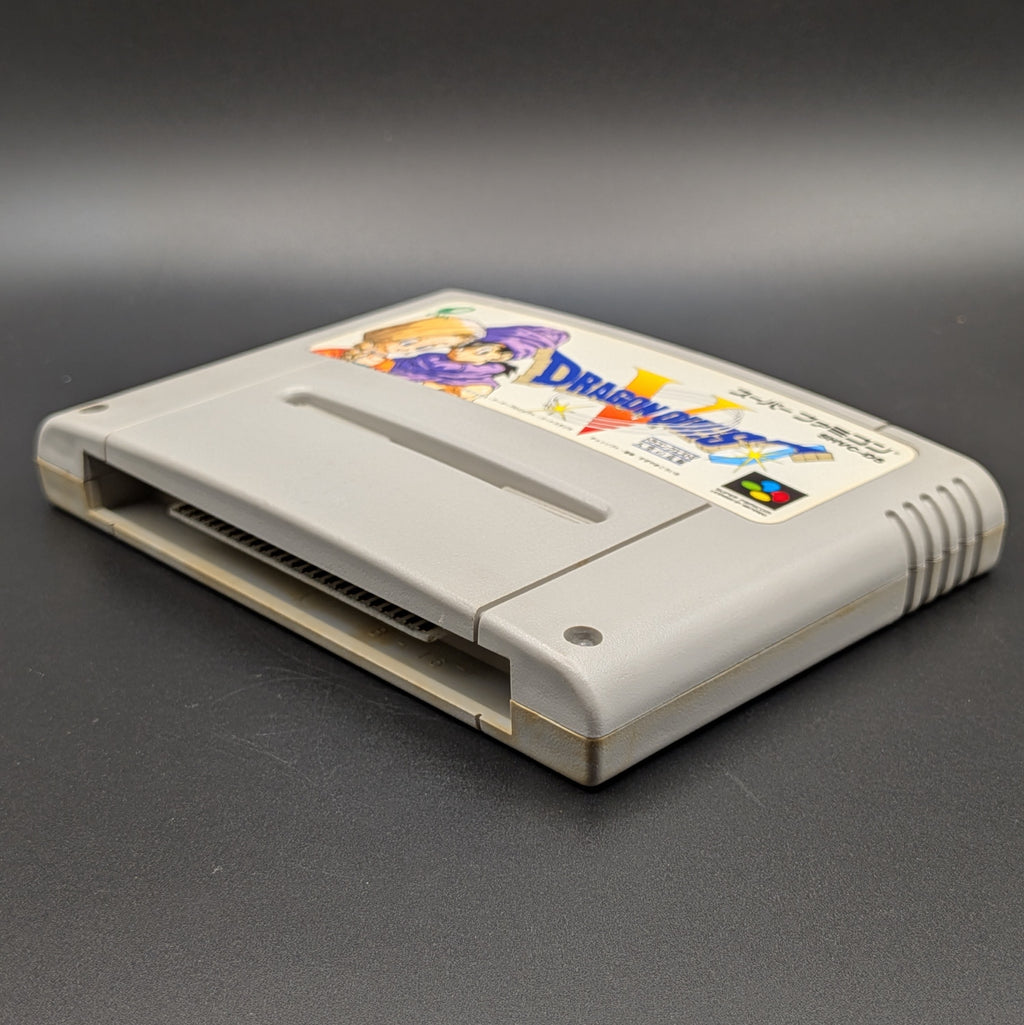Dragon Quest V video game cartridge for Nintendo's Super Famicom (SFC) Entertainment System, shown laying on its back with the label facing upwards. Showing the right hand side and bottom slot. It sits on a dark grey background.