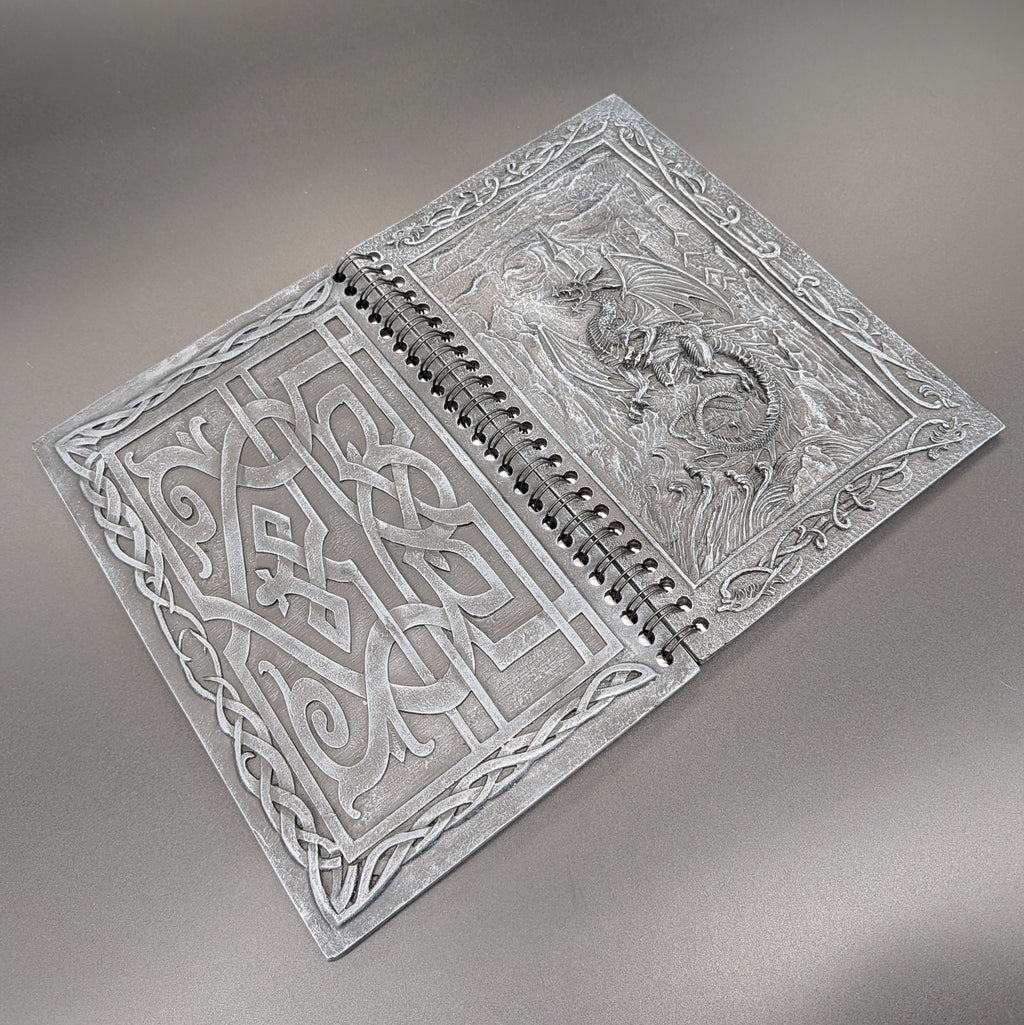 A spiral notebook with ornate Celtic knotwork and a dragon depicted on the cover, displayed on a neutral gray background.