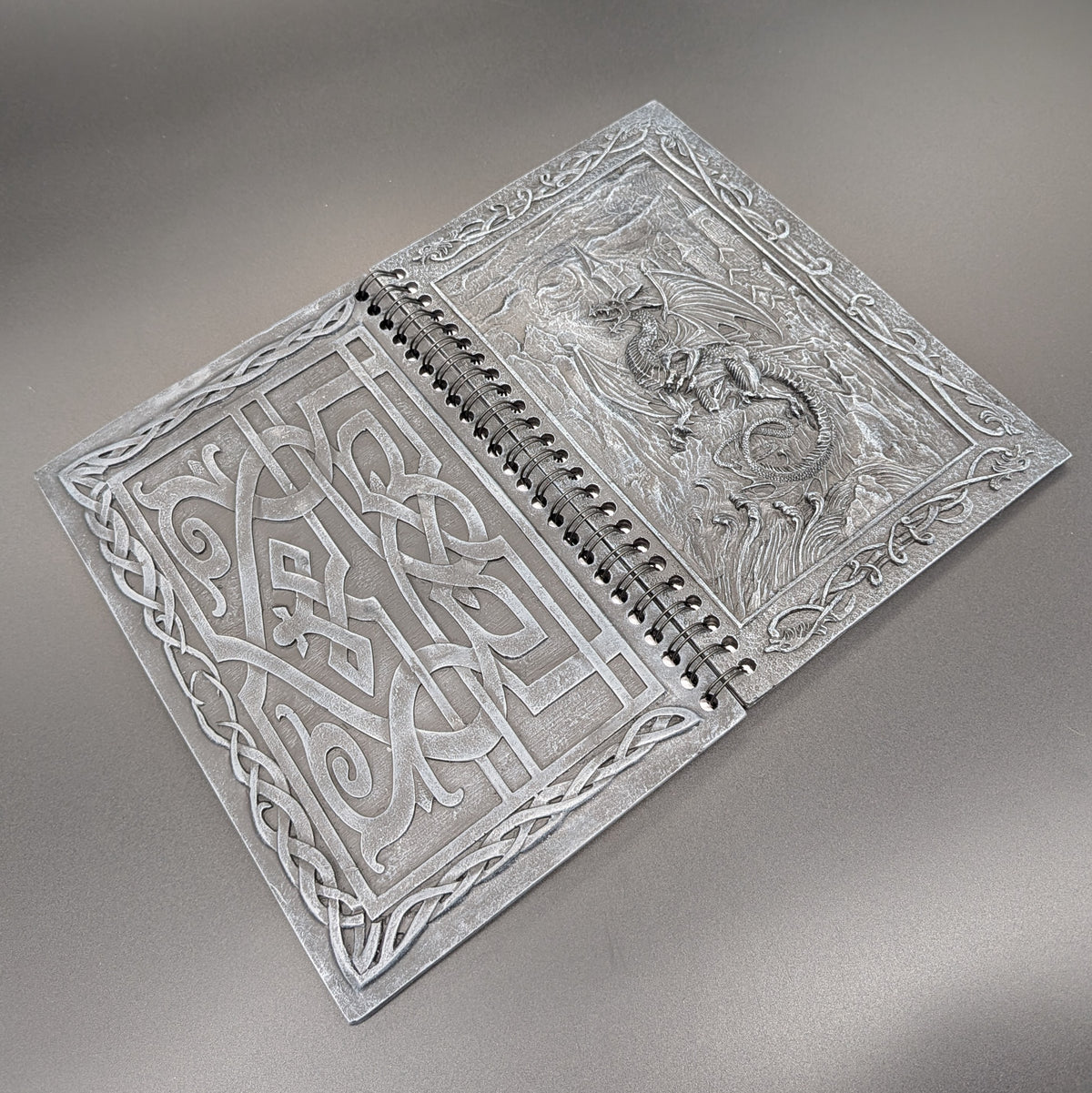 A spiral notebook with ornate Celtic knotwork and a dragon depicted on the cover, displayed on a neutral gray background.