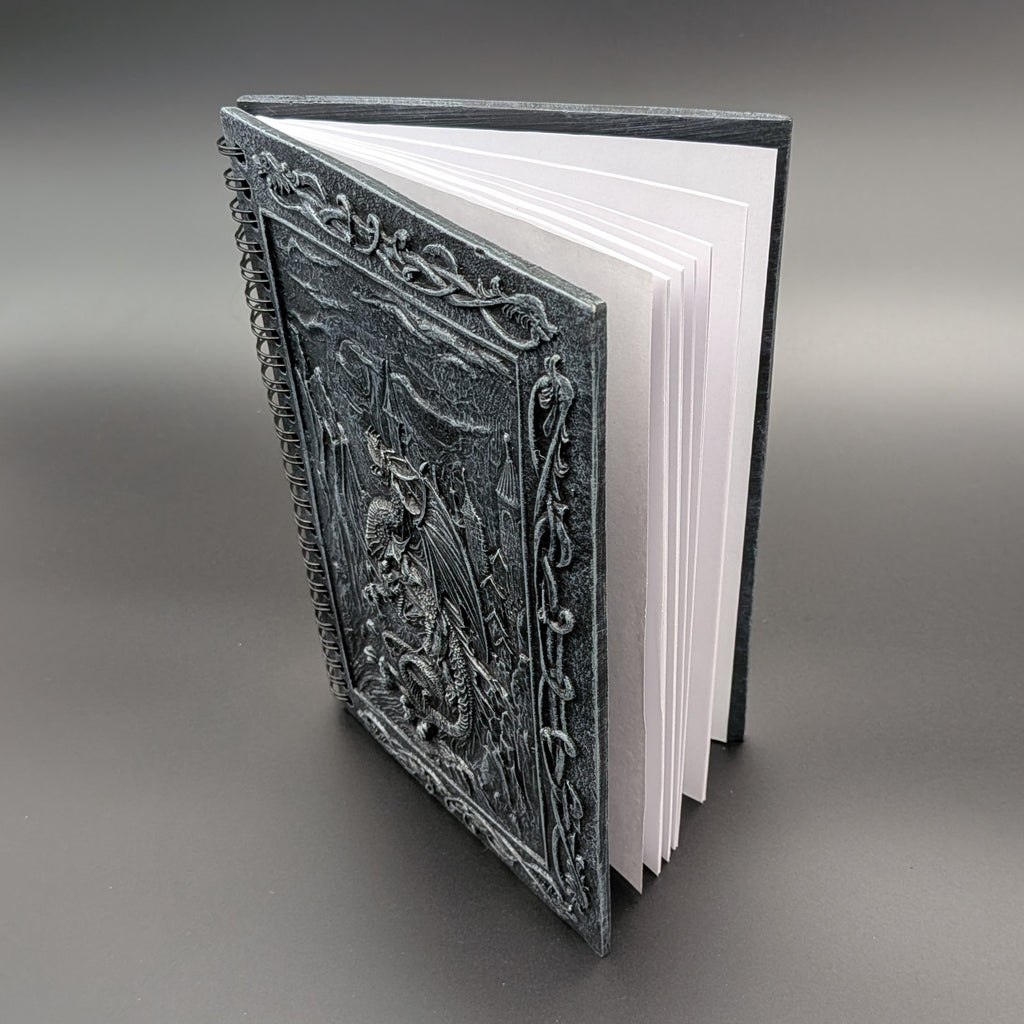 A hardcover book with an intricately cast dark gray cover featuring a dragon design, shown open to reveal white pages against a neutral gray background.