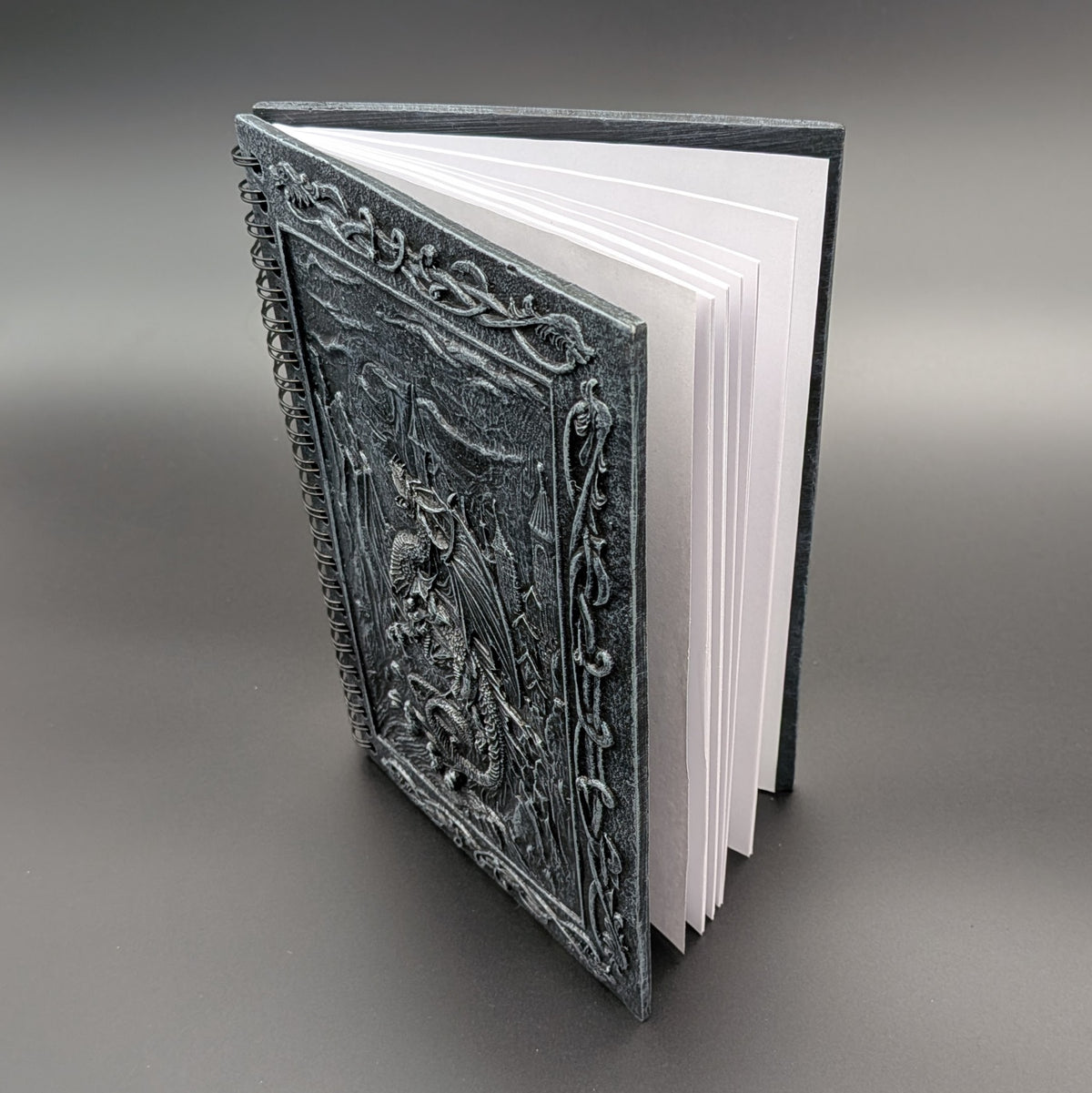 A hardcover book with an intricately cast dark gray cover featuring a dragon design, shown open to reveal white pages against a neutral gray background.