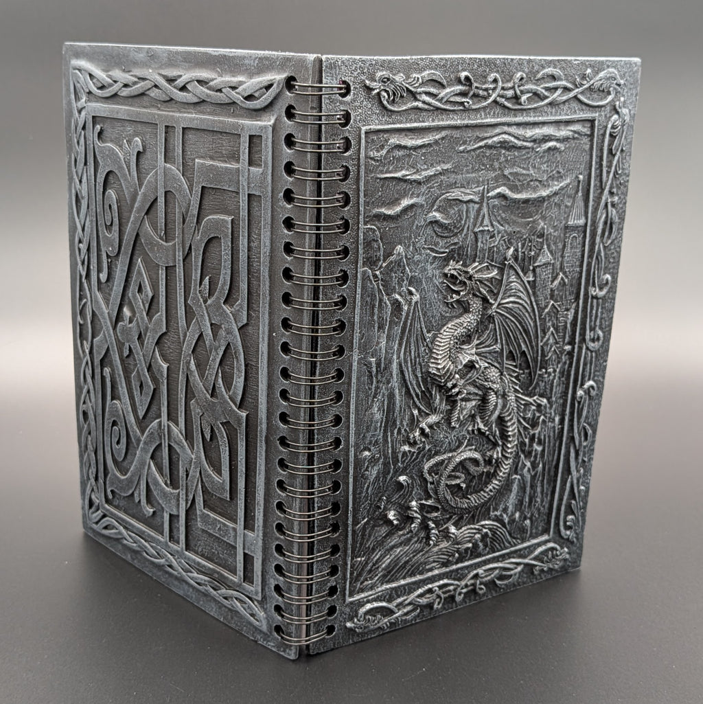A grey resin spiral notebook with Celtic knotwork patterns and a detailed dragon relief on the cover, photographed on a neutral gray background.
