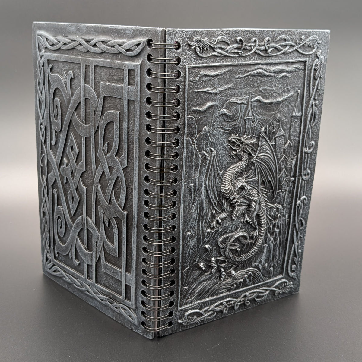 A grey resin spiral notebook with Celtic knotwork patterns and a detailed dragon relief on the cover, photographed on a neutral gray background.
