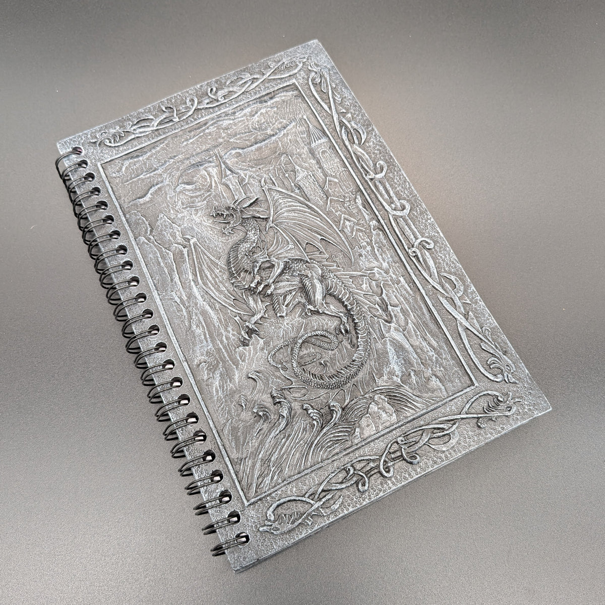 A spiral-bound notebook with an cast dragon design on the cover, photographed on a neutral gray background.