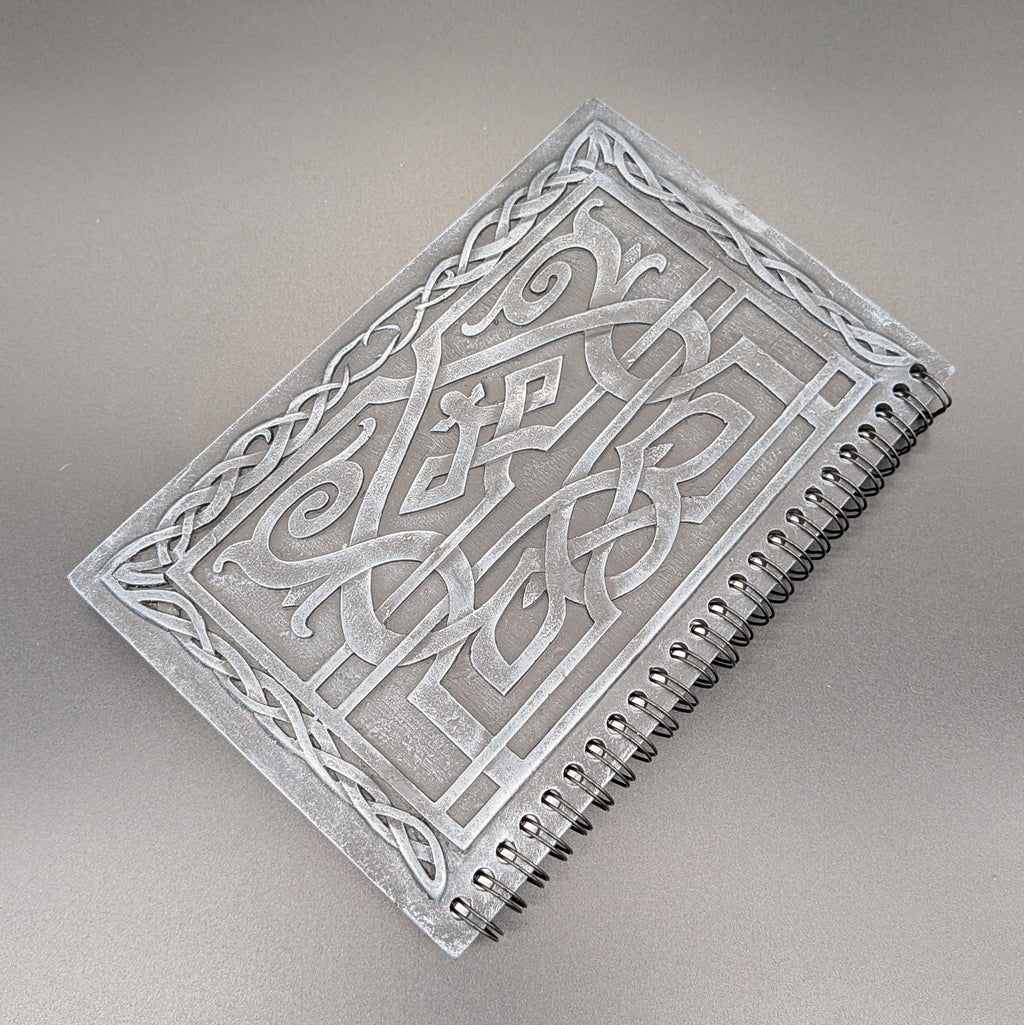 A spiral-bound notebook featuring a cast Celtic knot design, photographed on a neutral gray background.