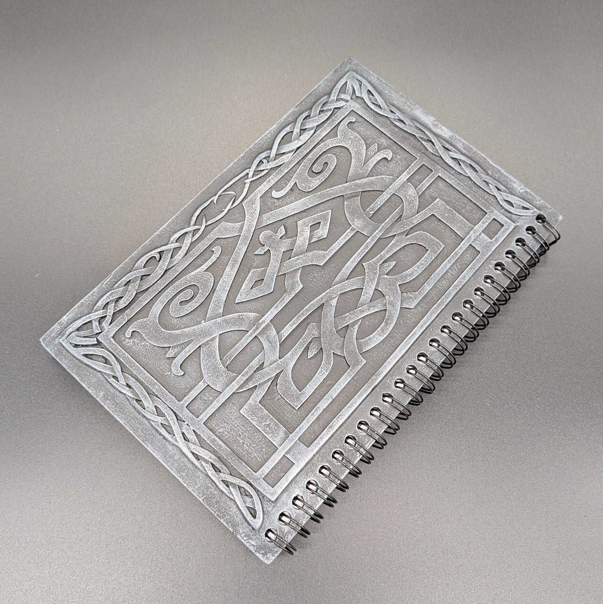 A spiral-bound notebook featuring a cast Celtic knot design, photographed on a neutral gray background.