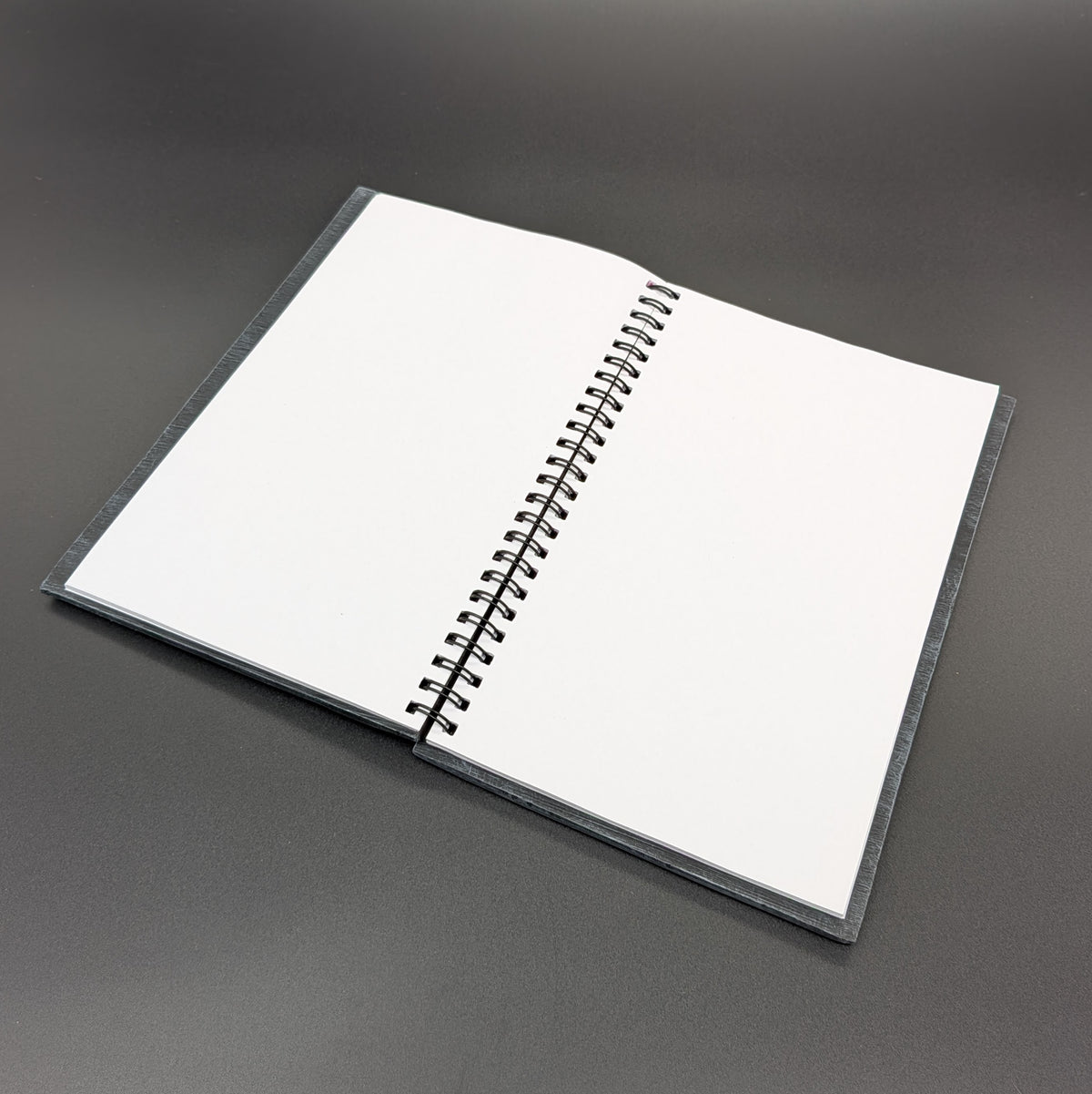 An open spiral-bound notebook with blank white pages, resting on a dark gray surface.