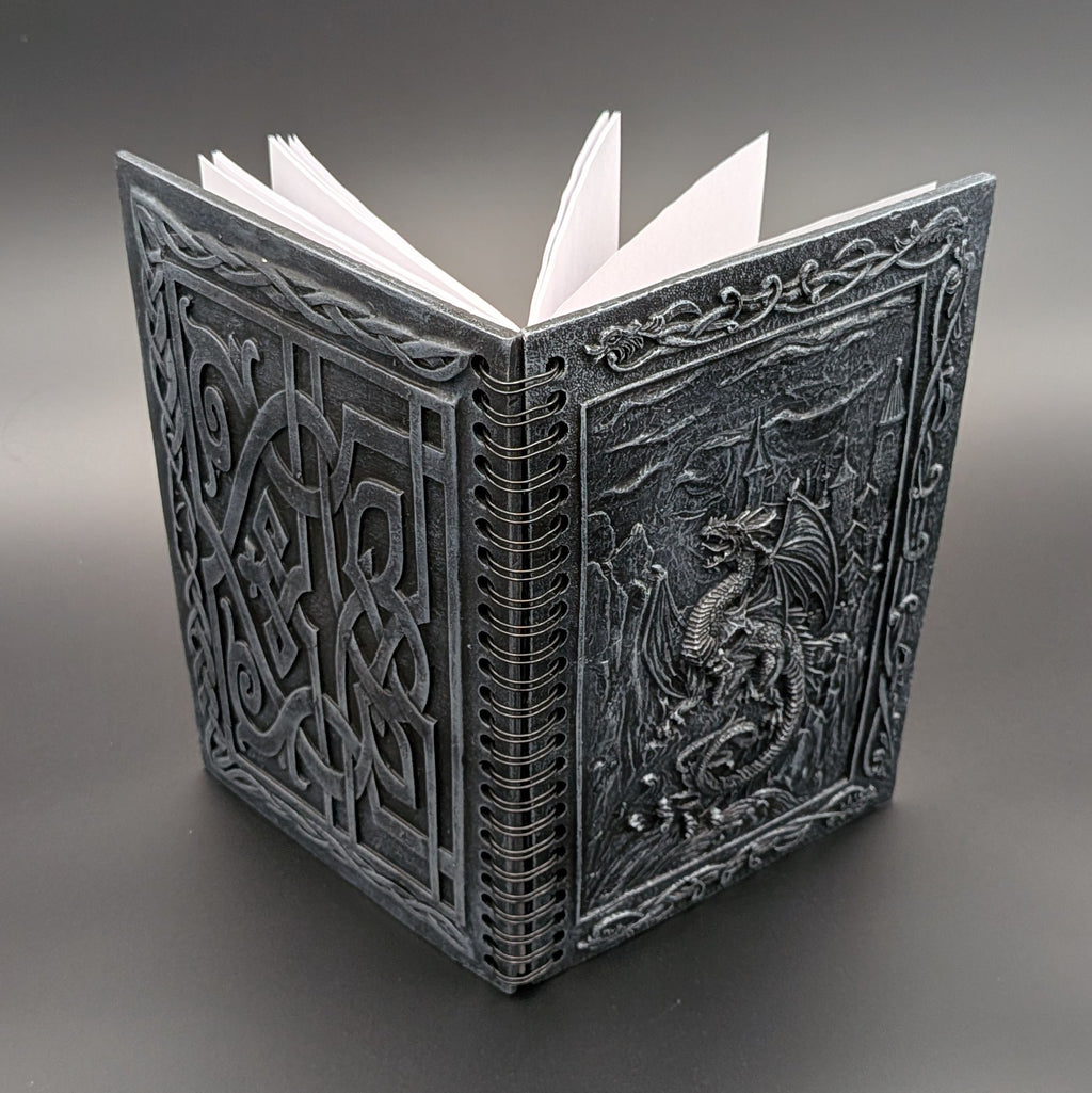 A dark gray resin notebook with white pages, featuring intricate Celtic knotwork on the back cover and a raised dragon design on the front cover, set against a dark gray gradient background.