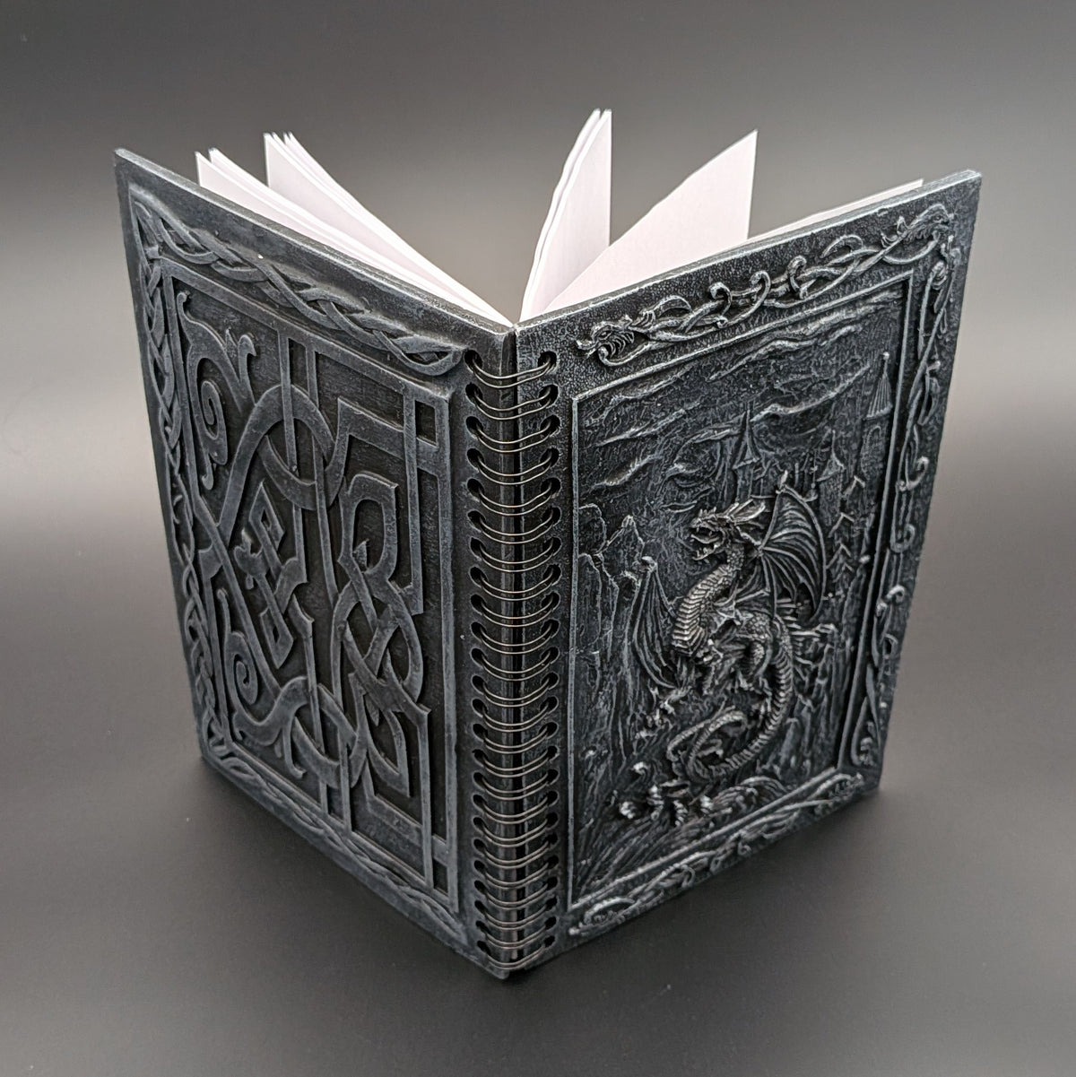 A dark gray resin notebook with white pages, featuring intricate Celtic knotwork on the back cover and a raised dragon design on the front cover, set against a dark gray gradient background.