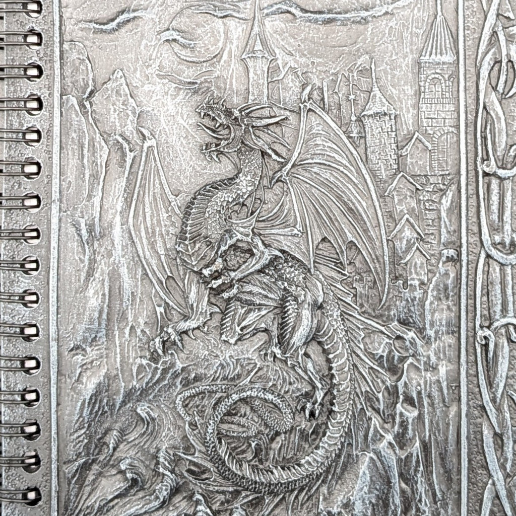 A close-up of a spiral-bound notebook cover featuring a cast fantasy dragon over a castle and mountain scene.