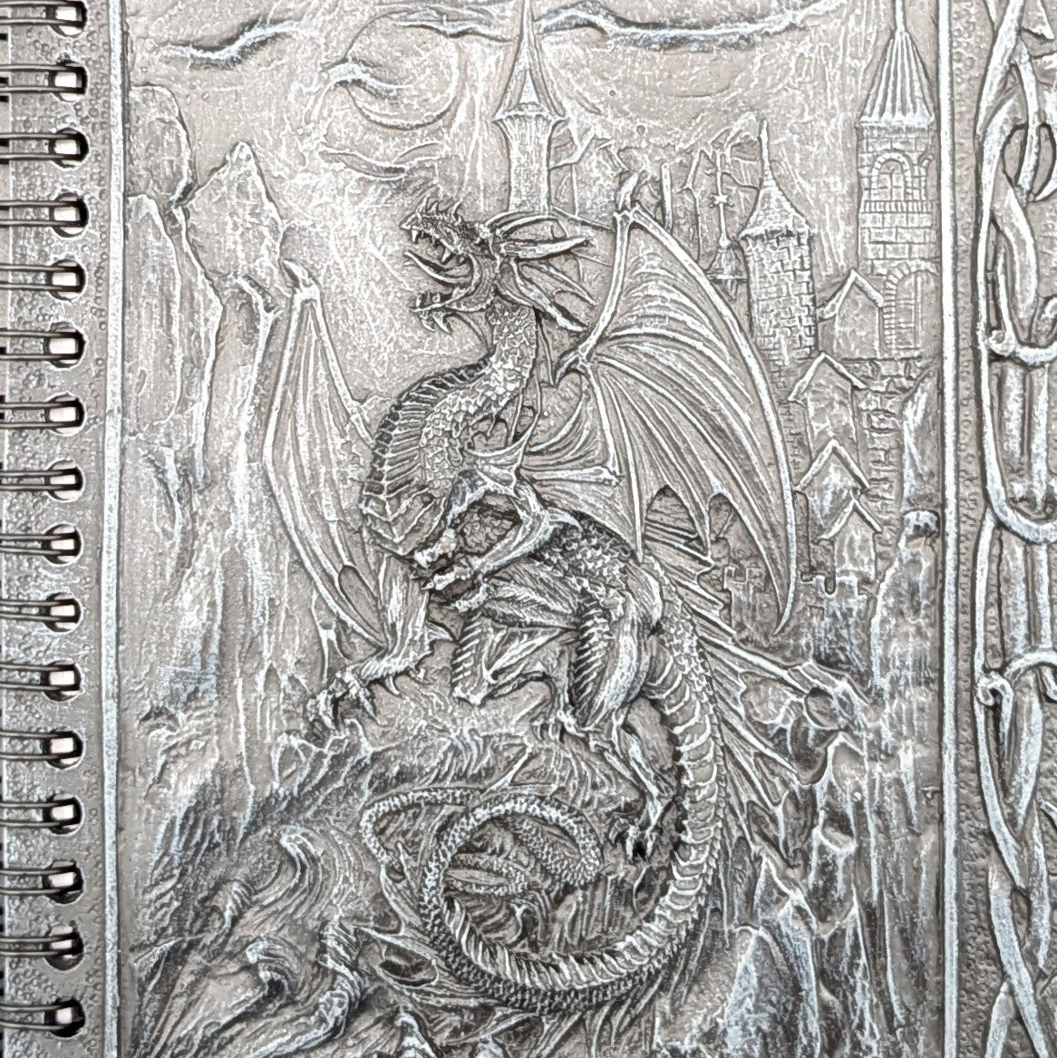 A close-up of a spiral-bound notebook cover featuring a cast fantasy dragon over a castle and mountain scene.