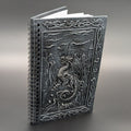A spiral-bound notebook with a dark gray cover featuring a detailed dragon and castle design, photographed on a neutral gray background.