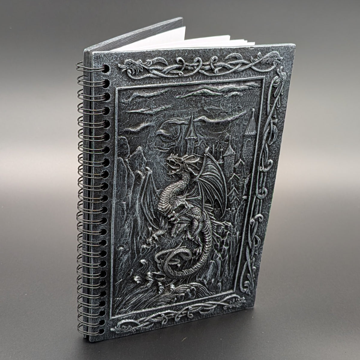 A spiral-bound notebook with a dark gray cover featuring a detailed dragon and castle design, photographed on a neutral gray background.