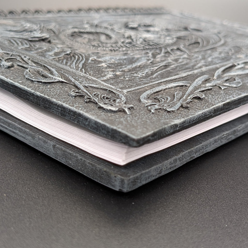 A close-up of a hardcover notebook with an ornate, cast resin dark gray cover featuring intricate design and scrollwork, resting on a dark surface.