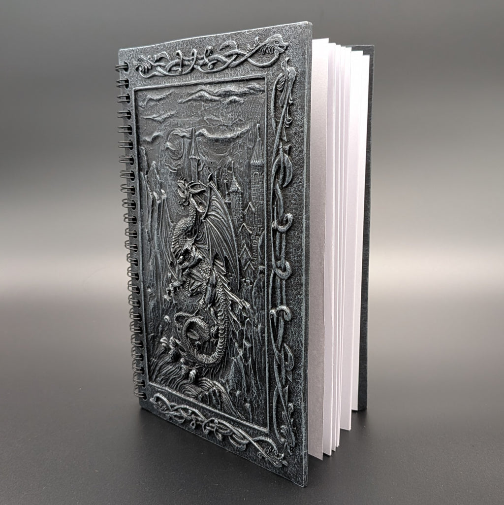 A spiral-bound notebook with a dark gray cast cover featuring a detailed dragon design, standing upright on a dark gray background.