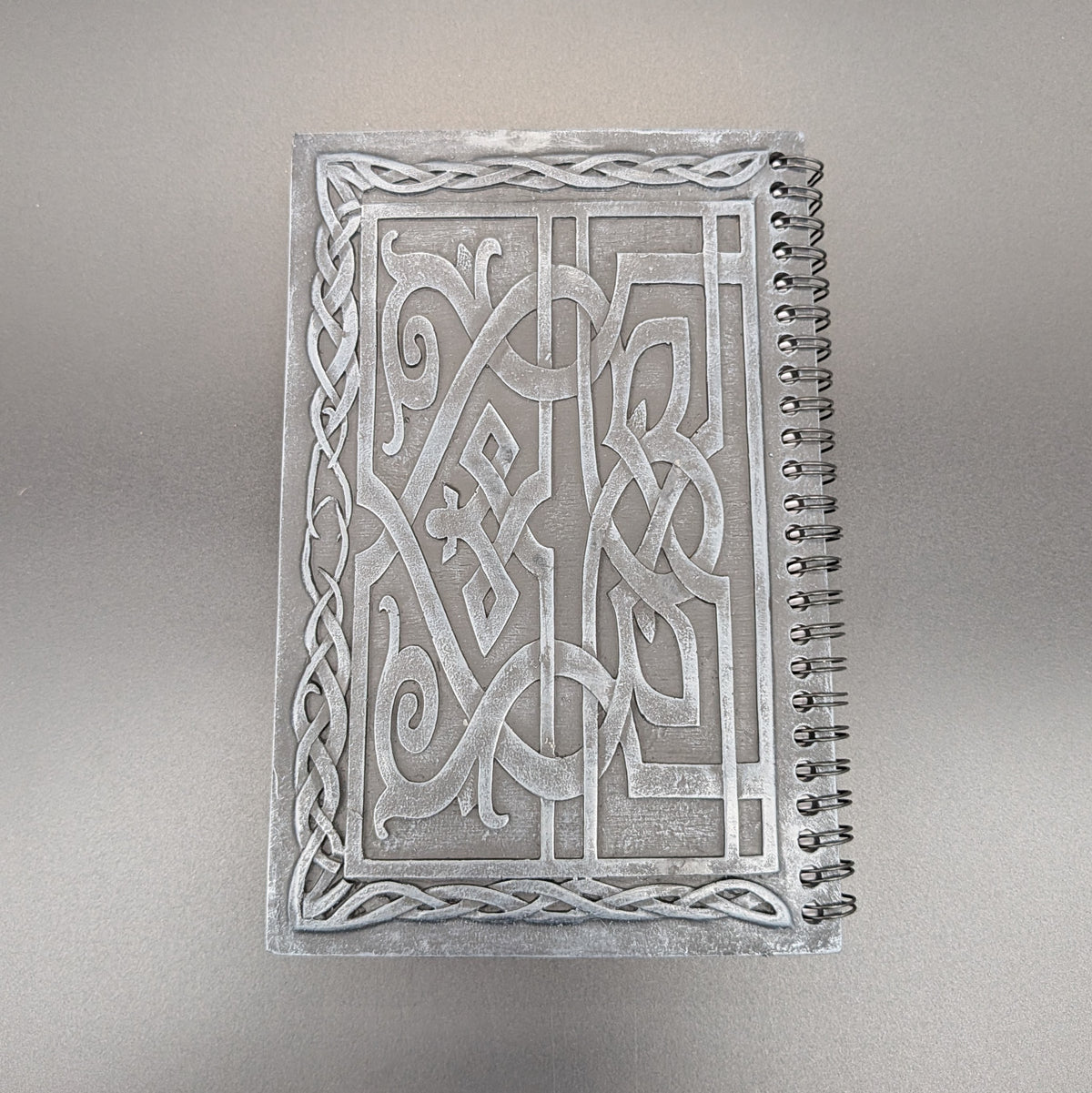 A spiral notebook with an cast Celtic knot design on the cover, photographed from directly above on a neutral gray background.