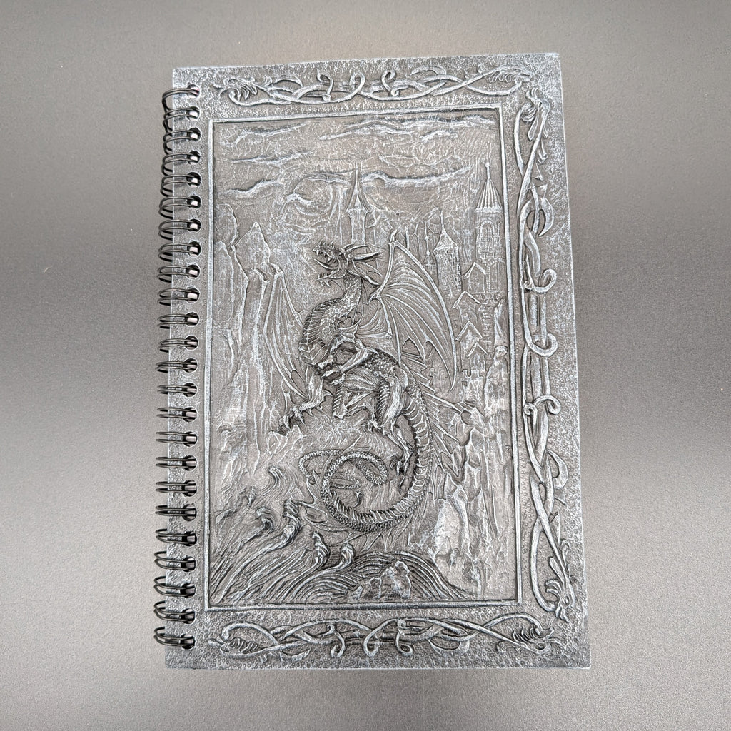 A spiral-bound notebook with a highly detailed cast dragon design on the cover, set against a neutral gray background.