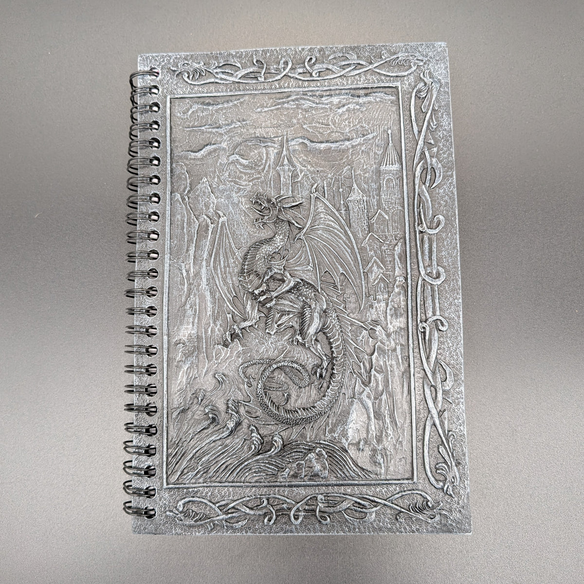 A spiral-bound notebook with a highly detailed cast dragon design on the cover, set against a neutral gray background.