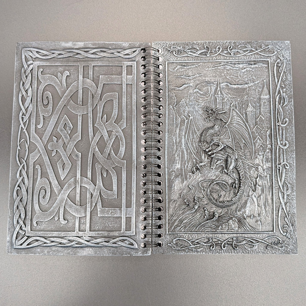 An open spiral-bound notebook with ornate cast covers featuring Celtic knotwork on the left page and a dragon scene on the right page, photographed against a plain light gray background.