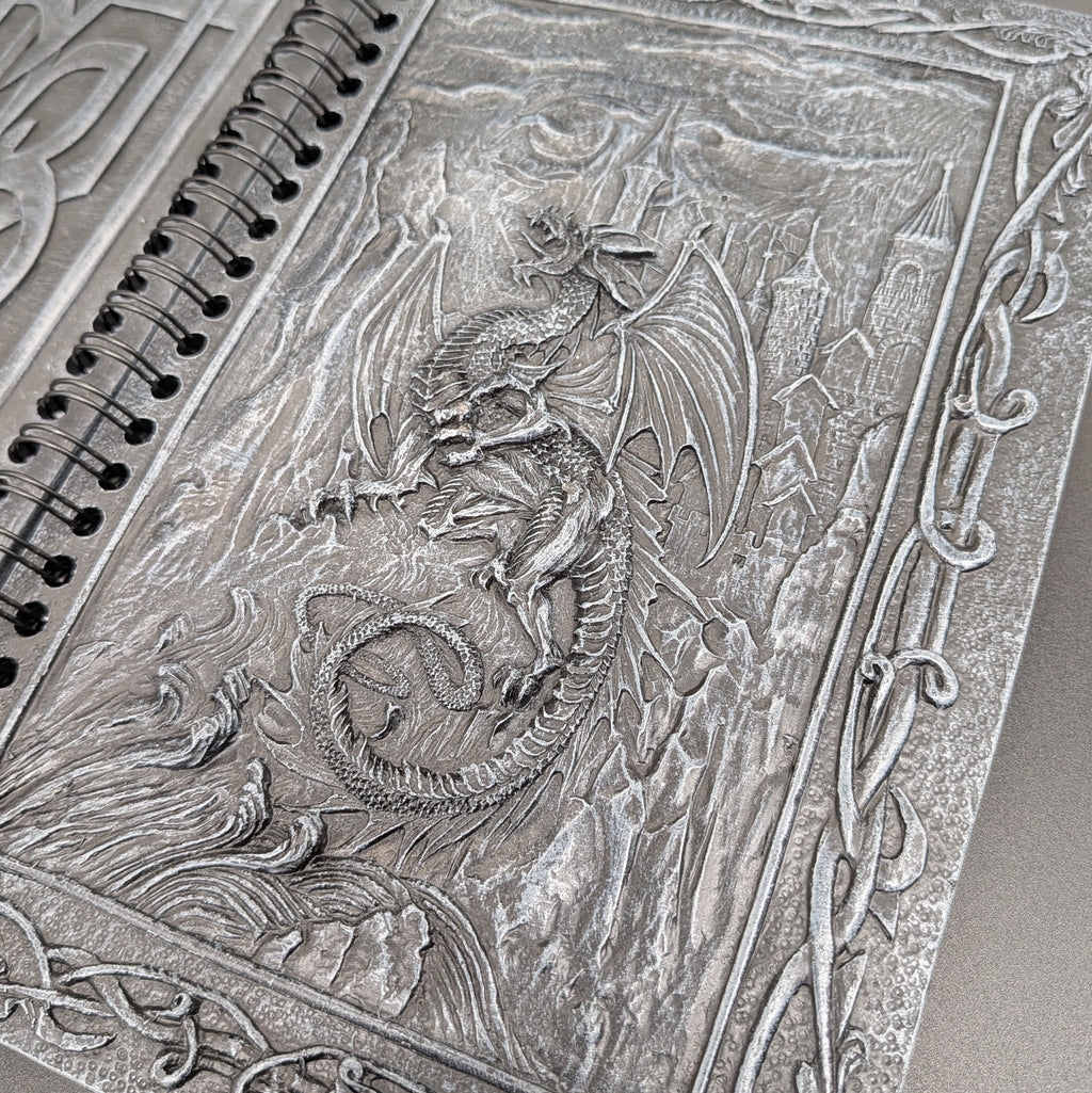 A close-up of a spiral notebook with an intricate dragon and castle cover design.