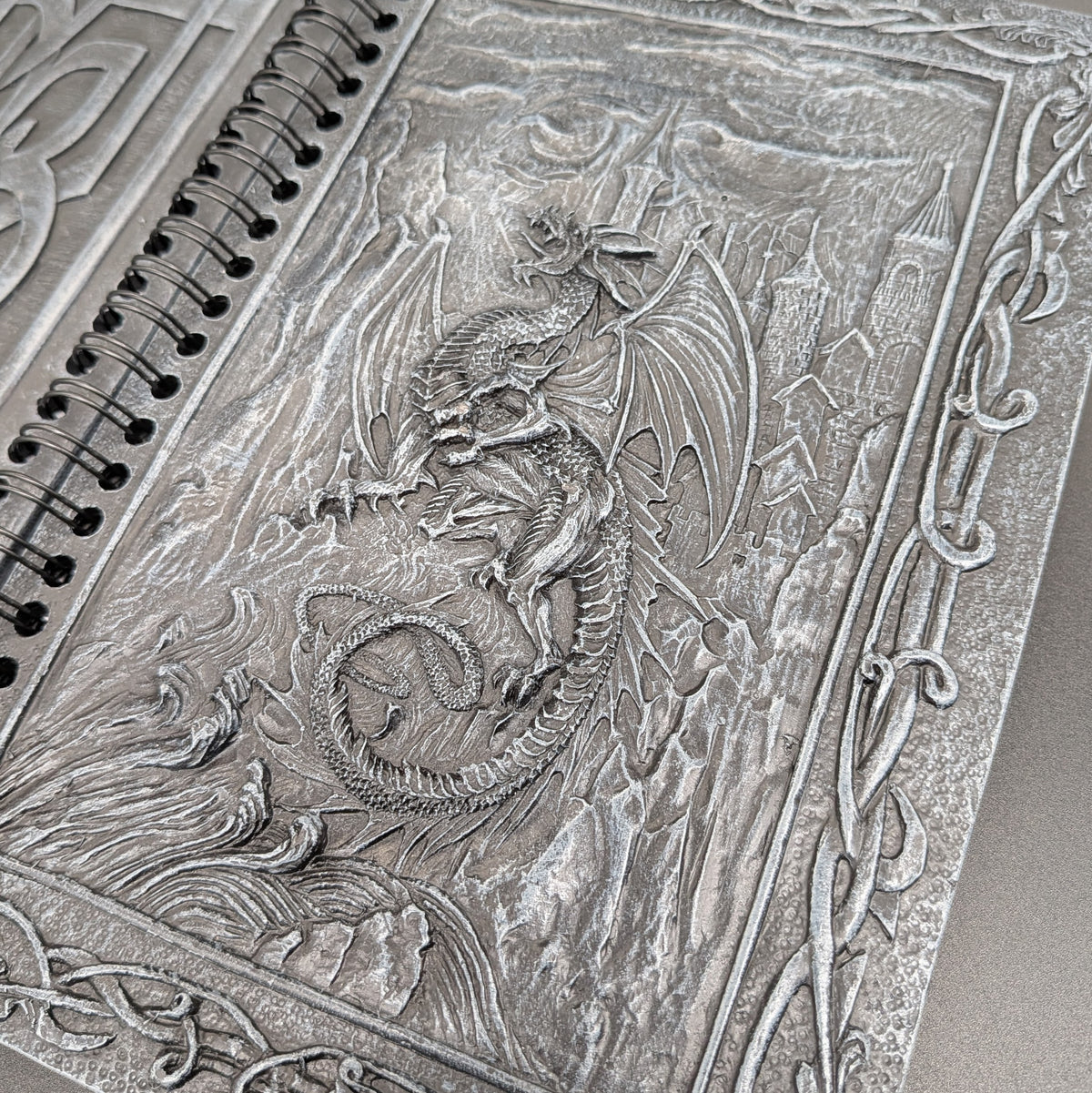 A close-up of a spiral notebook with an intricate dragon and castle cover design.