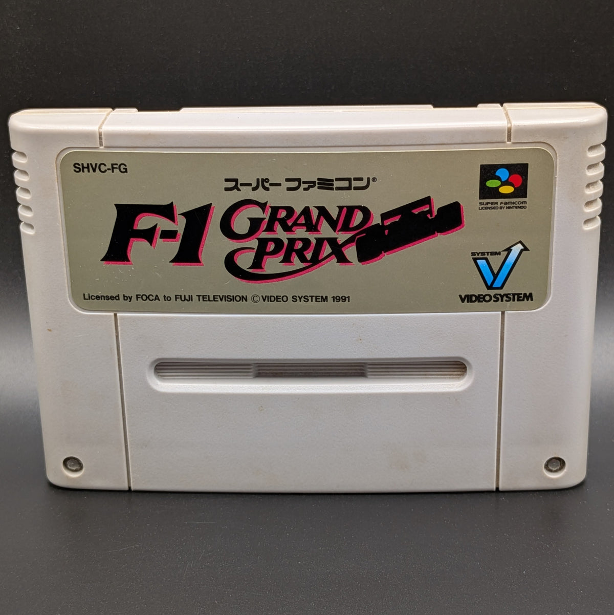 A close up of F-1 Grand Prix video game cartridge for Nintendo's Super Famicom (SFC) Entertainment System, shown standing up and facing forwards on a dark grey background.