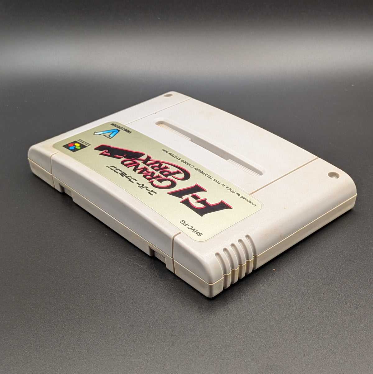 F-1 Grand Prix video game cartridge for Nintendo's Super Famicom (SFC) Entertainment System, shown laying on its back with the label facing upwards. Showing the left hand side and top edge. It sits on a dark grey background