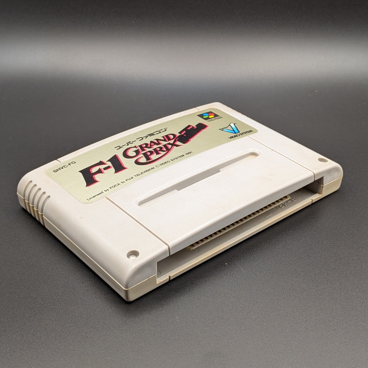 F-1 Grand Prix video game cartridge for Nintendo's Super Famicom (SFC) Entertainment System, shown laying on its back with the label facing upwards. Showing the left hand side and bottom slot. It sits on a dark grey background