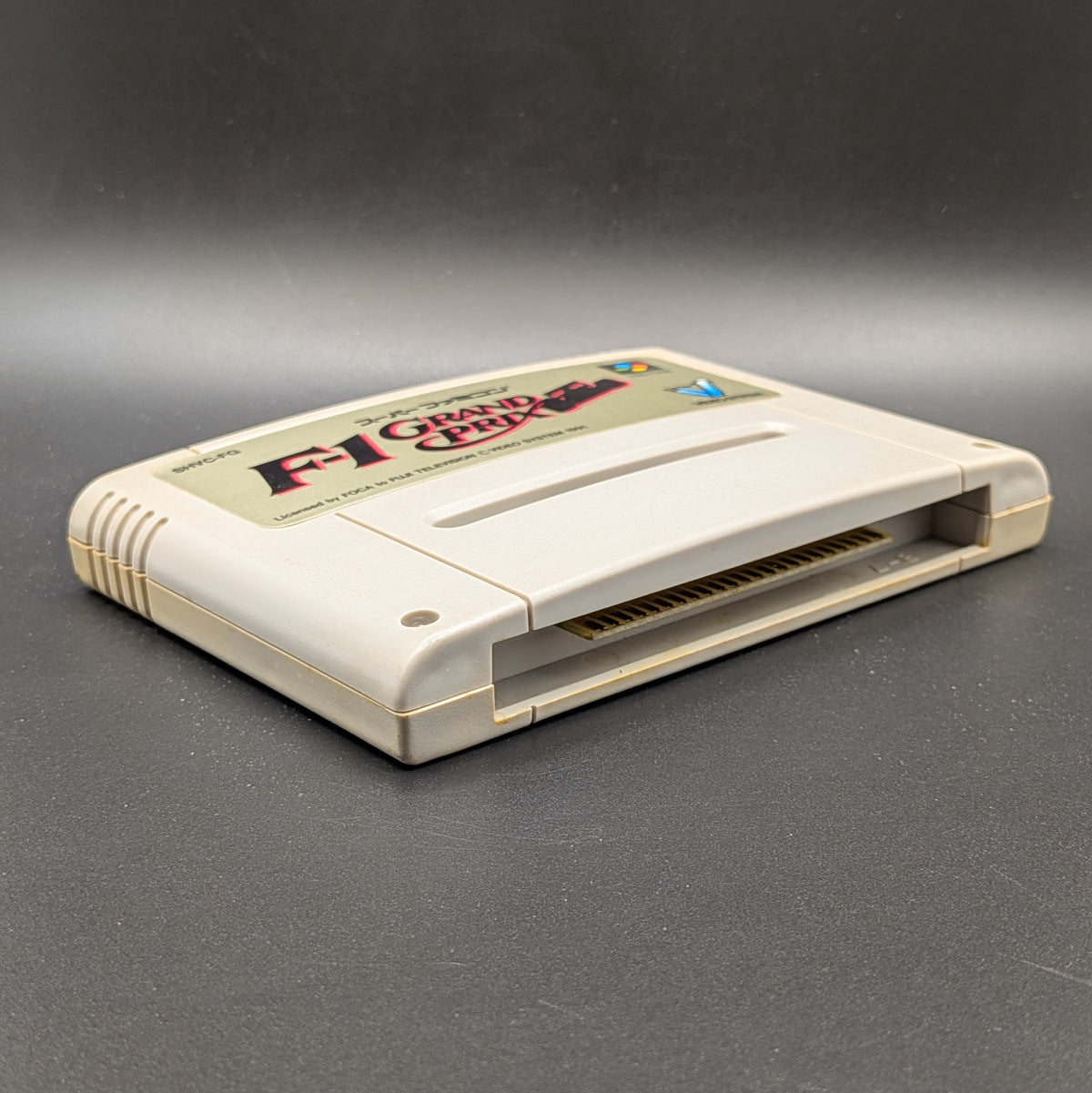 F-1 Grand Prix video game cartridge for Nintendo's Super Famicom (SFC) Entertainment System, shown laying on its back with the beige label facing up on a dark grey background.
