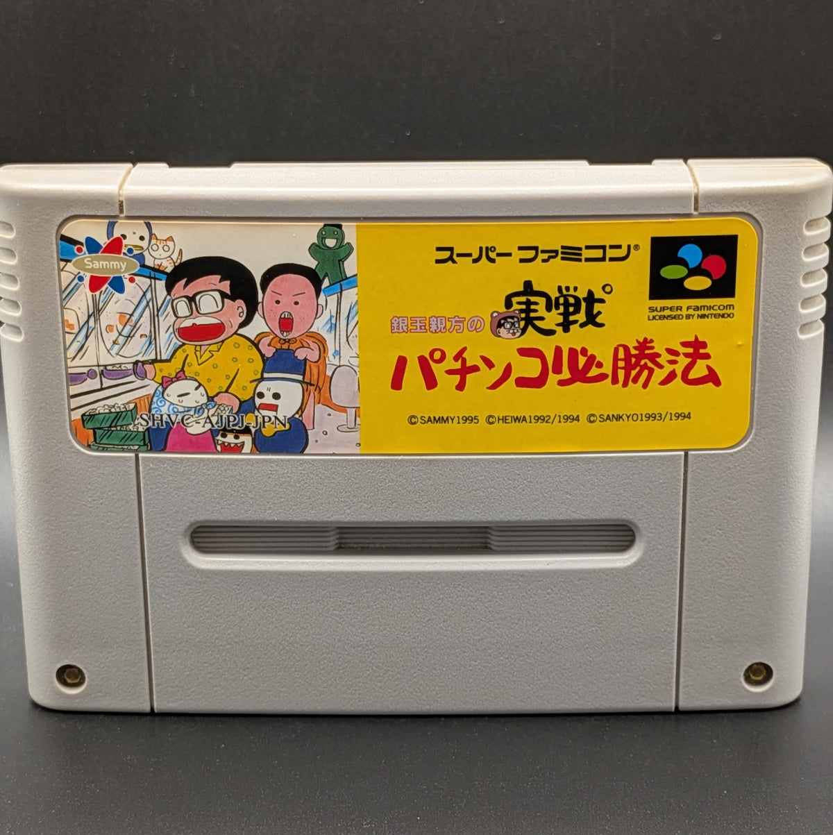 A Close up of Gindama Oyakata no Jissen Pachinko video game cartridge for Nintendo's Super Famicom (SFC) Entertainment System, shown standing up and facing forward on a dark grey background.
