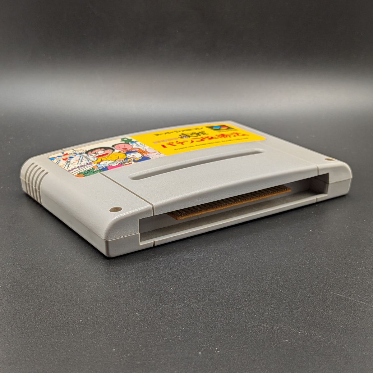 Gindama Oyakata no Jissen Pachinko video game cartridge for Nintendo's Super Famicom (SFC) Entertainment System, shown laying on its back, label facing up on a dark grey background.
