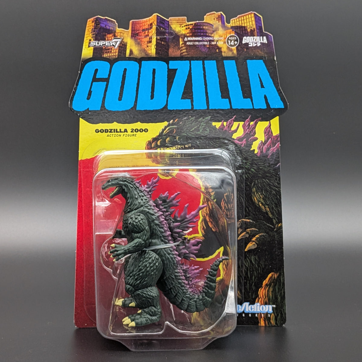 Godzilla 2000 Action Figure in its original carded blister packaging, featuring a dark green textured figure with pink spines and yellow claws, set against a dark gray background.