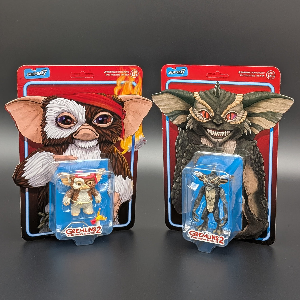 Two Gremlins 2 action figure packages displayed side by side on a dark gray surface. The left package features the Baby Gremlin figure, and the right package features the Big Gremlin figure, both in their original blister packaging with red backing cards.