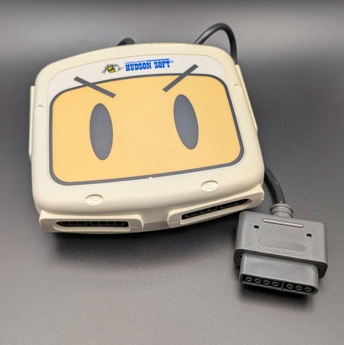 A vintage Hudson Soft interface device with a beige plastic casing, a yellow face graphic, and an attached gray connector cable, shown on a dark gray background.