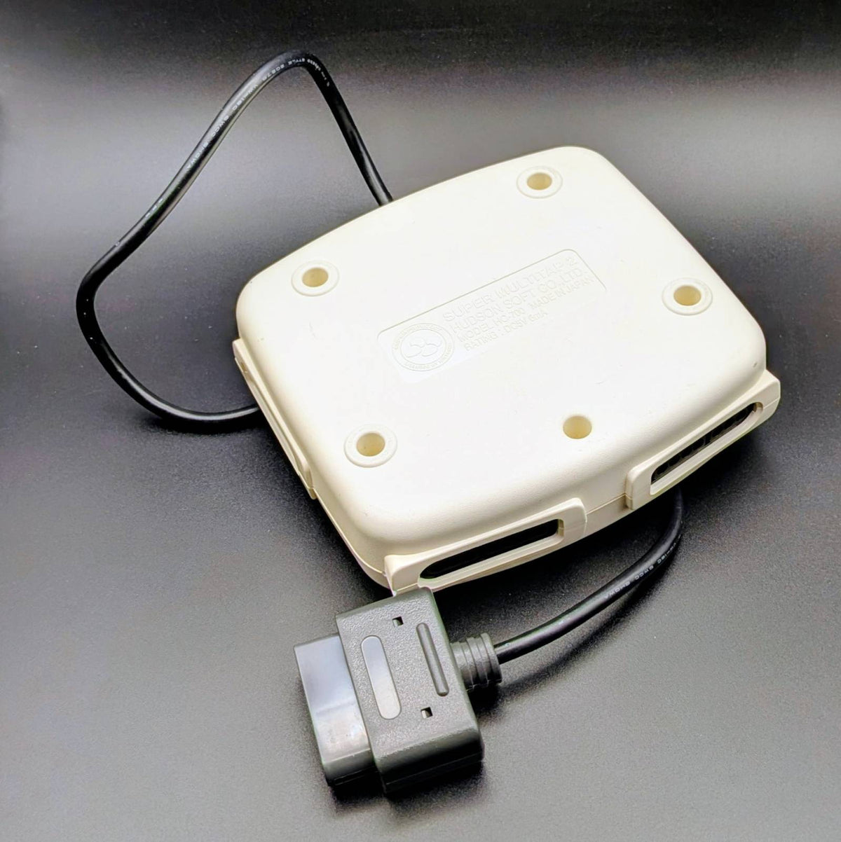 A vintage beige plastic electronic device with a black power cable and connector, photographed on a dark gray surface.