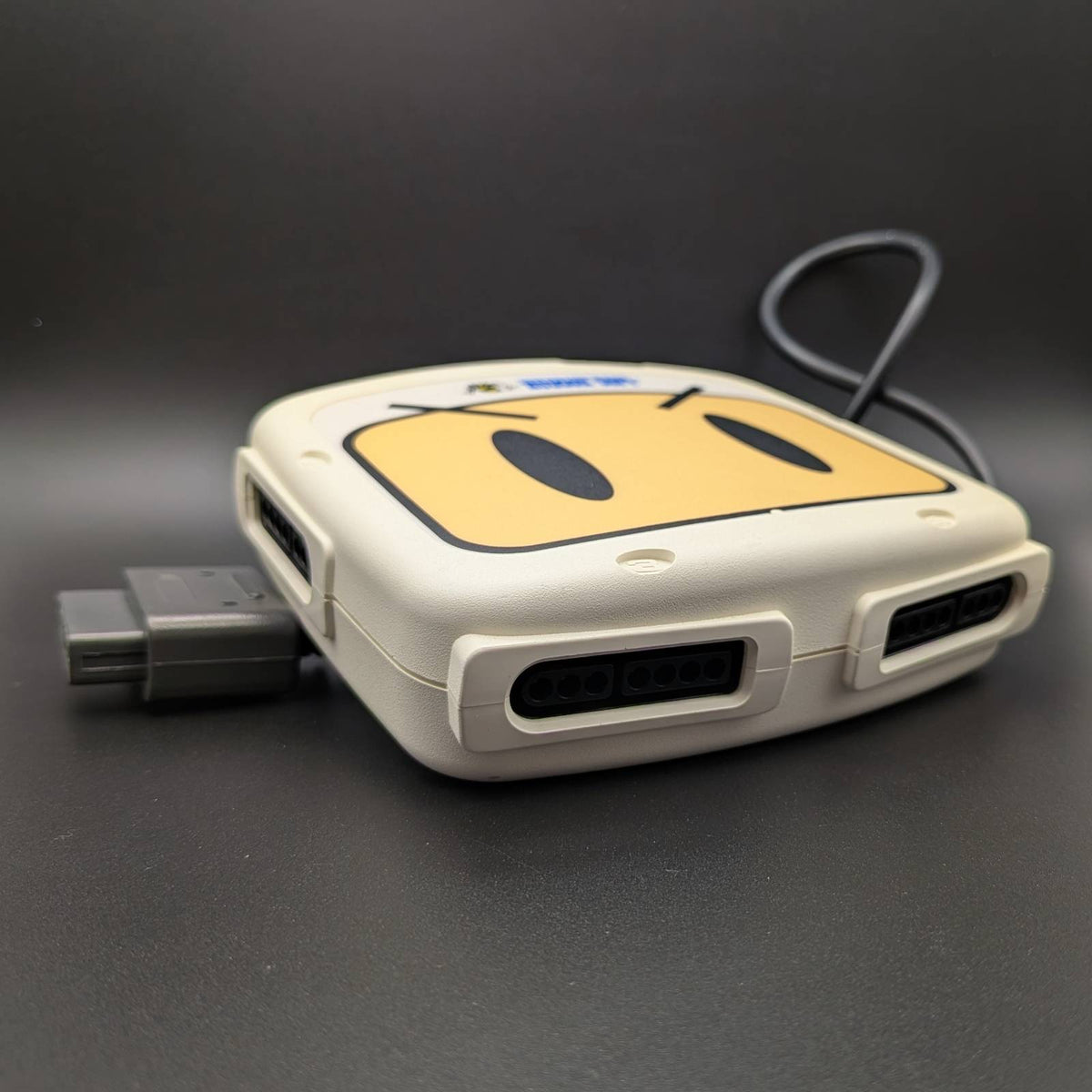 A white and yellow retro-style hub with a cartoon face graphic on top, shown with a cable plugged in on a dark gray background.