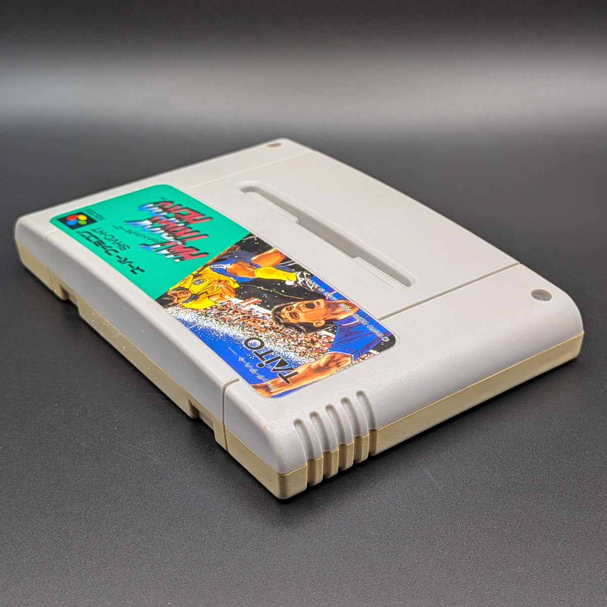 Hat Trick Hero video game cartridge for Nintendo's Super Famicom (SFC) Entertainment System, shown laying on its back with the label facing upwards. Showing the left hand side and top edge. It is displayed on a dark grey background.