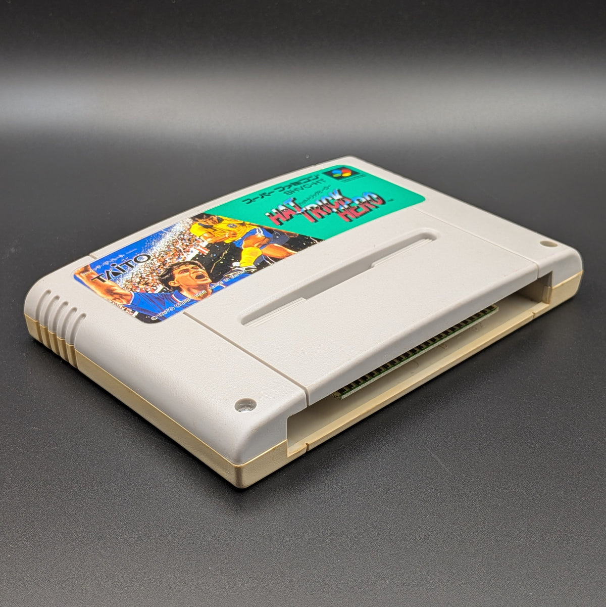 Hat Trick Hero video game cartridge for Nintendo's Super Famicom (SFC) Entertainment System, shown laying on its back with the label facing upwards. Showing the left hand side and bottom slot. It is displayed on a dark grey background.