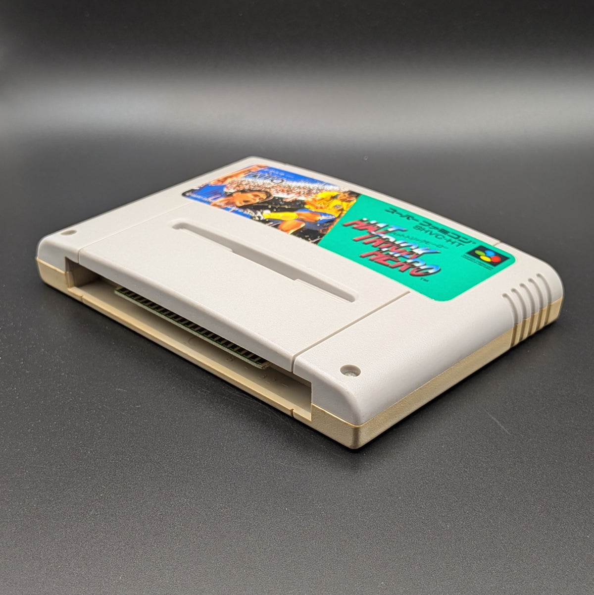 Hat Trick Hero video game cartridge for Nintendo's Super Famicom (SFC) Entertainment System, shown laying on its back with the label facing upwards. Showing the right hand side and bottom slot. It is displayed on a dark grey background.