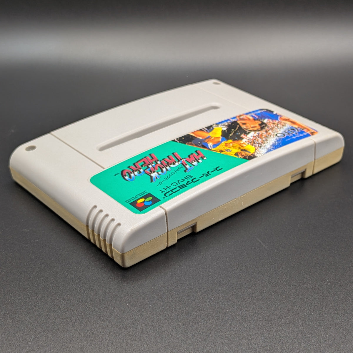 Hat Trick Hero vvideo game cartridge for Nintendo's Super Famicom (SFC) Entertainment System, shown laying on its back with the label facing upwards. Showing the right hand side and top edge. It is displayed on a dark grey background.